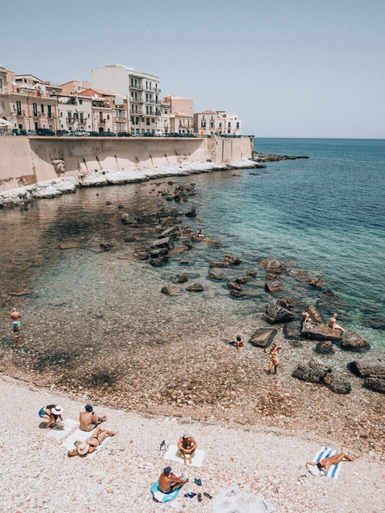 Rocky beach with swimmers along a Sicilian coastline lined with pastel buildings.