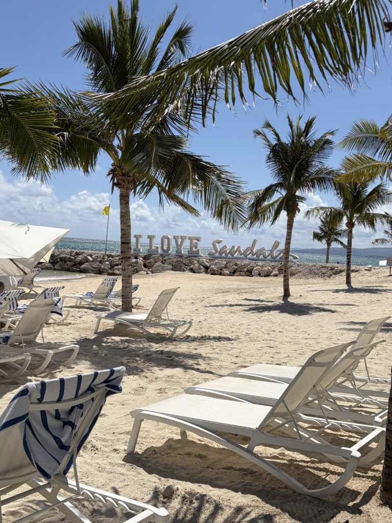 Beachfront view at Sandals Dunn’s River featuring the “I Love Sandals” sign set against the turquoise Caribbean water and sandy beach in Jamaica.