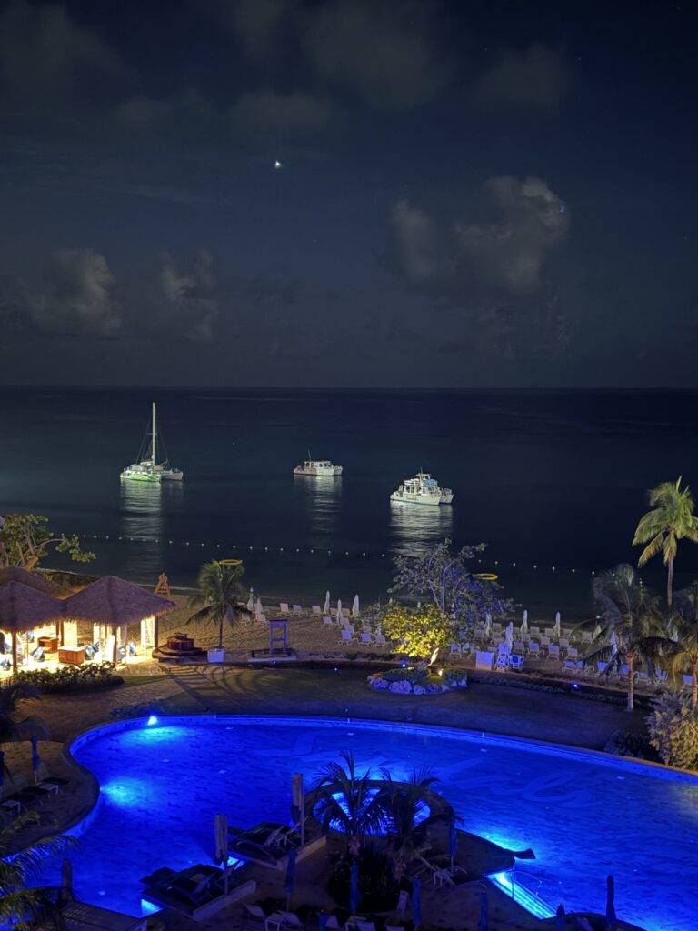 Nighttime view from the Ocarina rooftop bar at Sandals Dunn’s River overlooking the resort and the water, with three illuminated boats in the distance and a relaxed tropical atmosphere.