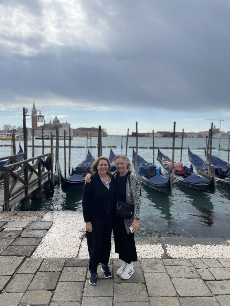 Two sisters standing in front of traditional gondolas along a canal in Venice, Italy.